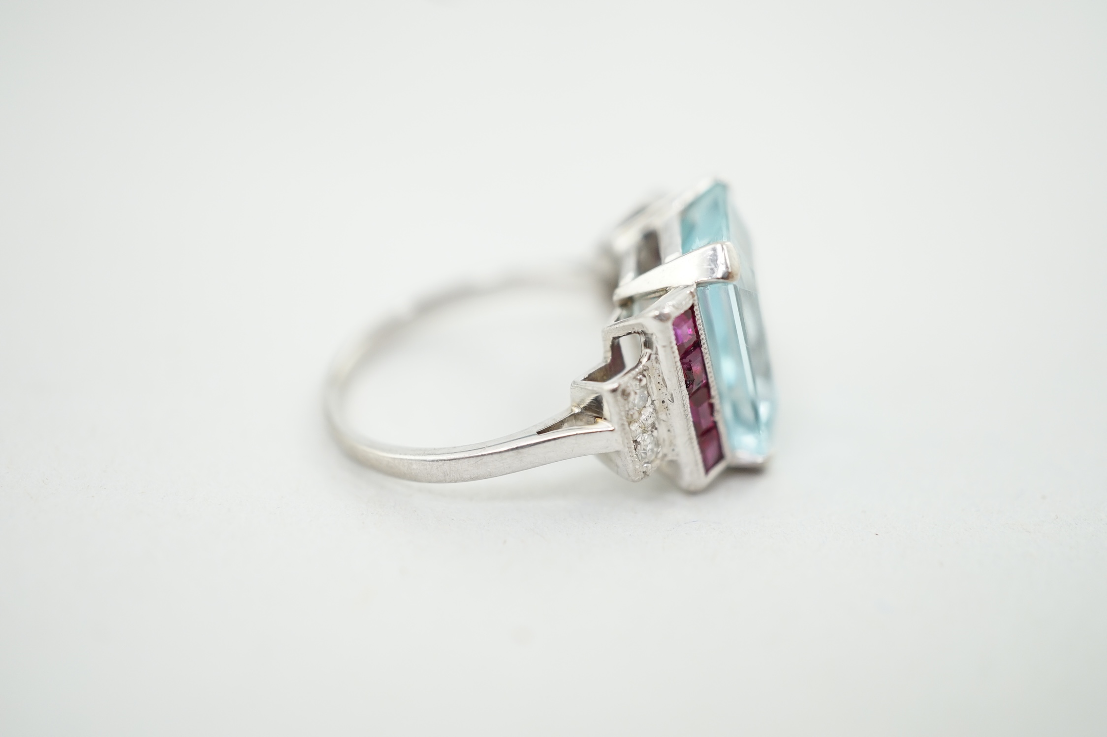 An Art Deco style platinum? and single stone emerald cut aquamarine set dress ring, with millegrain set ruby and diamond cluster stepped shoulders